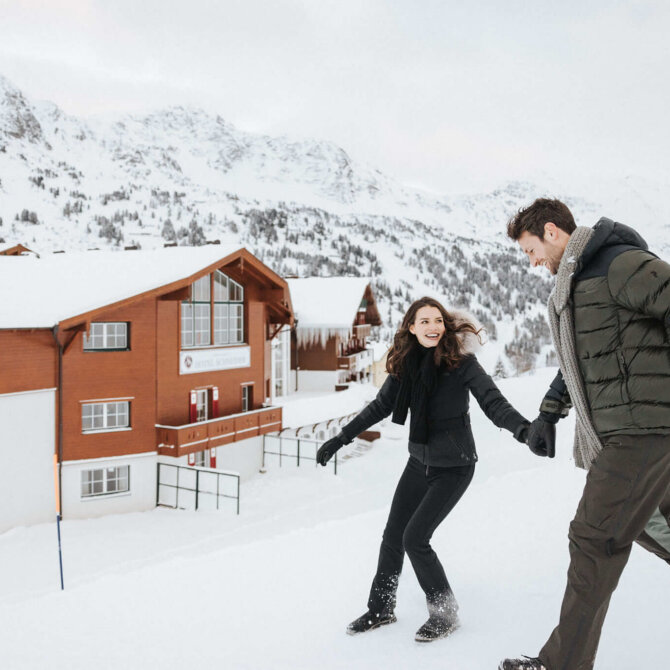 Paar im Schnee im Hintergrund das Hotel Schneider